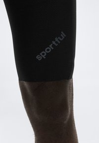 Κοντινή λήψη μαύρου υφασμάτινου κολάν αθλητικής ένδυσης με την λέξη "Sportful" τυπωμένη διαγώνια στο μηρό πάνω σε σκούρο δέρμα.