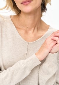 Kobieta w beżowym swetrze z długim rękawem, z delikatnie splecionymi rękami przy piersi, częściowo widoczną twarzą i szyją.