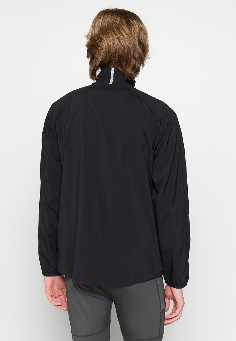 Persoon met kort bruin haar draagt een zwart langetermijnjacket en grijze broek, staand met de rug naar de camera tegen een plain witte achtergrond.