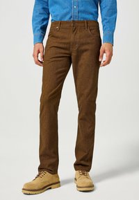 Pantalones marrones texturizados con un corte recto, que cuentan con un cierre frontal de botón y cremallera, combinados con botas de ante beige claro con cordones.