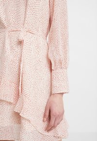 Robe longue à manches longues rose clair avec de petits pois rouges, ceinture à la taille, ourlet à volants et une main reposant sur le côté.