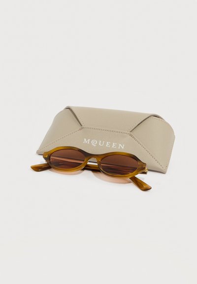 Καφέ οβάλ γυαλιά ηλίου με κεχριμπαρένιο σκελετό που βρίσκονται μπροστά από μία μπεζ δερμάτινη θήκη προστασίας με ανάγλυφο το λογότυπο McQueen.