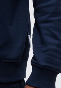 Κοντινό πλάνο στα μανίκια ενός ναυτικού μπλε φούτερ με ορατή ετικέτα της μάρκας στη πλαϊνή ραφή και ριμπ μανσέτες στους καρπούς.