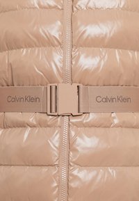 Bežová prešívaná bunda s lesklým povrchom, so zipsom a látkovým opaskom s logom "Calvin Klein" v zladenom odtieni v strede.
