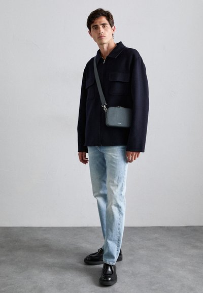 Mörk marinblå zip-up jacka med två framfickor, ljusblå raka jeans och svarta snörskor. Liten grå axelremsväska.