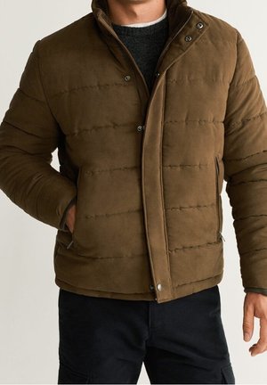 Homme portant une veste matelassée marron avec fermeture éclair et boutons-pression, pantalon noir et un pull sombre en dessous, debout avec une main dans la poche.