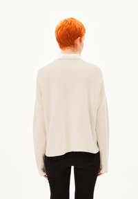 Helle beige Bluse mit langen Ärmeln im lockeren Schnitt, ausgestattet mit einem runden Ausschnitt und einem leicht verkürzten Saum. Über schwarzen Hosen getragen.