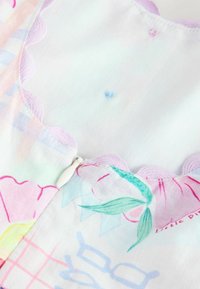 Primo piano di un capo color pastello con stampa floreale, bordo rosa smerlato e piccolo tirazip bianco al collo.