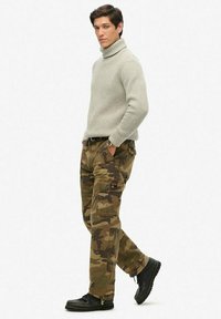 Jersey de cuello alto a rayas grises, pantalones cargo de camuflaje con múltiples bolsillos y botas negras con cordones. Materiales texturizados con una paleta de colores combinada.