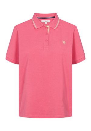 Femme portant un polo rose à manches courtes avec un petit logo et un jean bleu, debout devant un fond uni.
