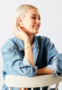 Bracciale di perline con accenti verdi, bianchi e gialli indossato al polso. Il modello indossa una giacca di jeans e si siede su una sedia bianca.