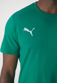 Zeleni bombažni T-majica s belim logotipom Puma na levem prsnem delu. Standardna okrogla ovratnica in kratki rokavi z gladko teksturo.