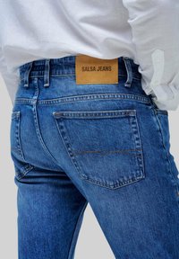 Vaqueros de mezclilla azul con un corte ajustado, con una etiqueta de cuero que dice "SALSA JEANS", bolsillos traseros visibles y detalles de costura en contraste.