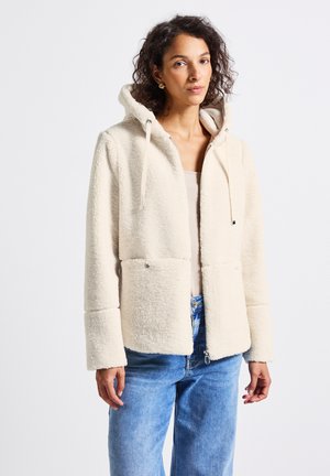 Frau trägt eine cremefarbene Fleece-Zip-Up-Softshelljacke mit Kordelzügen und blaue Jeans vor weißem Hintergrund.