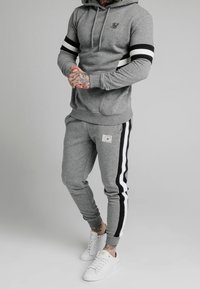 Mężczyzna w szarym dresie z kapturem i spodniach dresowych w czarno-białe paski, w białych sneakersach, stojący na jednolitym tle.