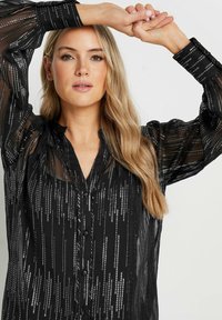 Blusa nera con una trama trasparente e linee verticali argento, abbottonatura frontale, vestibilità comoda con maniche a sbuffo e colletto.