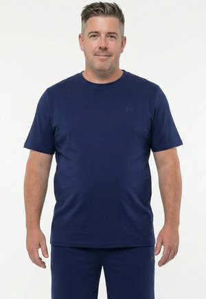 Lyhyttukkainen mies, jolla on yksivärinen tummansininen t-paita ja matching-housut, seisoo valkoista taustaa vasten.