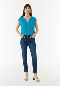 Türkise Bluse mit kurzen Ärmeln und V-Ausschnitt, kombiniert mit dunkelblauen Slim-Fit-Jeans und hellen Blockabsatzsandalen. Schlichtes Design.