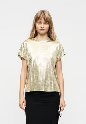 Mlada žena s dugom svijetlosmeđom kosom, odjevena u sjajnu zlatnu majicu kratkih rukava i crnu suknju, stoji pred jednobojnom svijetlom pozadinom.