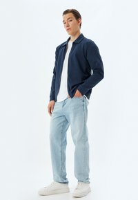 Chaqueta de cremallera navy sobre una camisa blanca, combinada con jeans de color azul claro y zapatillas blancas. Tejido suave con un corte relajado y cuello.