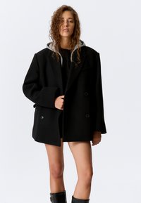 Cappotto nero doppiopetto con vestibilità comoda, realizzato in lana. Presenta un cappuccio grigio, grandi bottoni e maniche dritte.