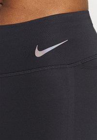 Leggings atléticos negros con una textura suave, cintura alta y un logo reflectante de Nike en la cadera izquierda. Detalles cosidos a lo largo de las costuras.