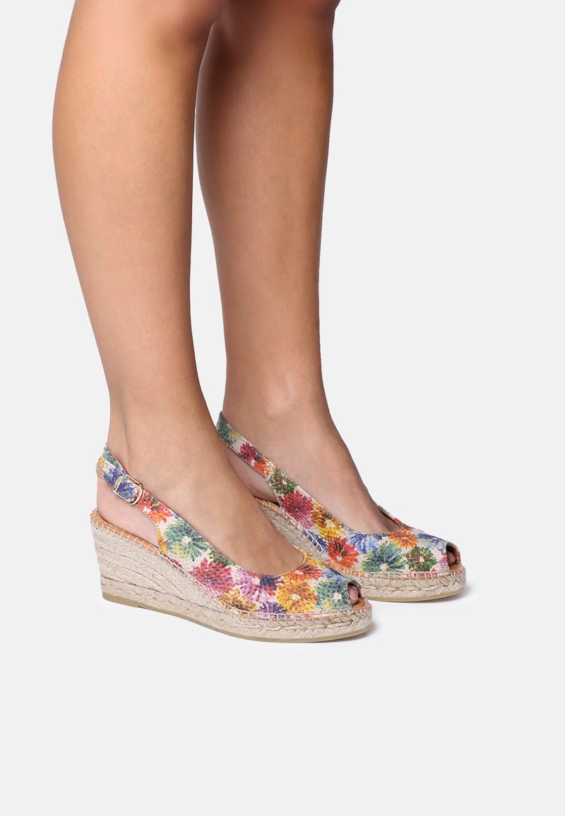Benen met kleurrijke wedge espadrille sandalen van bloemrijk stof, met open tenen en enkelbandjes, op een lichte achtergrond.