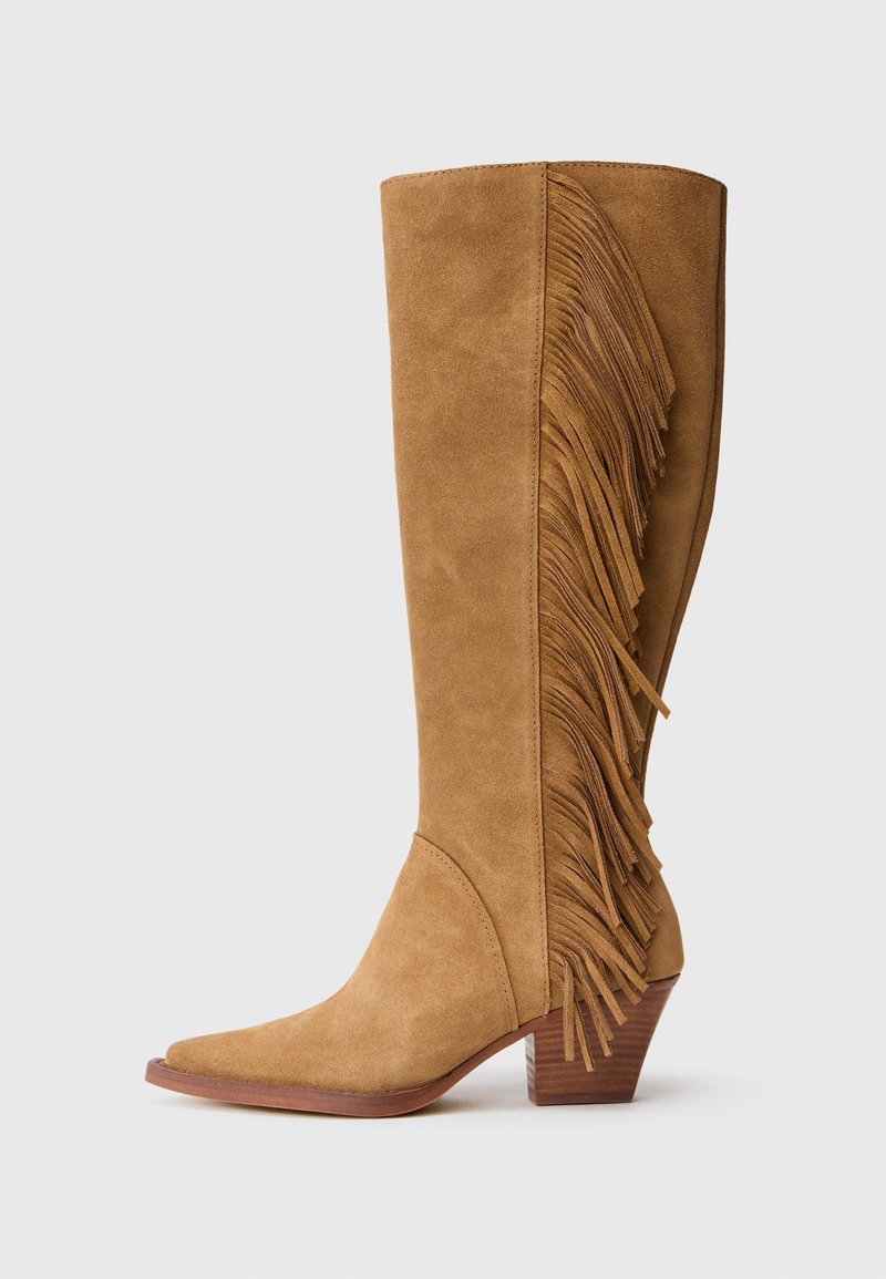 Botas de ante en color marrón claro, de caña alta, con punta afilada, que presentan flecos verticales en el costado y un tacón de madera apilada.