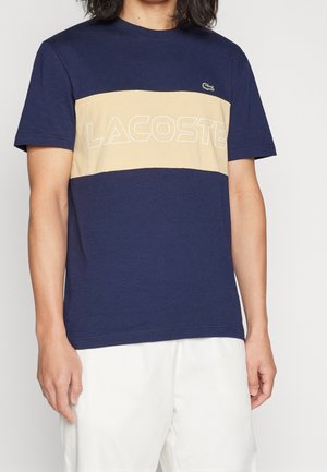Blue cotton t-shirt with a cream horizontal center band featuring "LACOSTE" text. Small green logo on the left chest. Short sleeves.