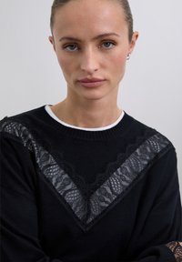 Jeune femme avec les cheveux tirés en arrière portant un pull noir orné d'accents en dentelle en forme de V et d'un col blanc, sur fond uni.