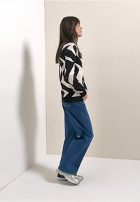 Trui met zwart-wit abstract patroon, gecombineerd met blauwe wijde broeken. Model draagt zilveren schoenen. Eenvoudige achtergrond.