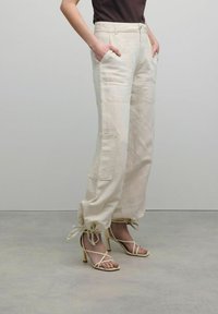 Vrouw in beige cargobroek met vastgebonden cuffs en beige sandaaltjes met hoge hakken, handen in de zakken, staande op een grijze vloer.