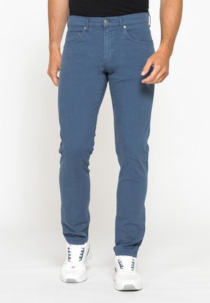 Uomo con pantaloni slim fit blu e sneakers bianche in piedi su uno sfondo semplice.