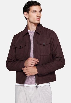 Young man looking right wearing a dark brown zip jacket over a light purple sweater and light-colored pants against plain background.