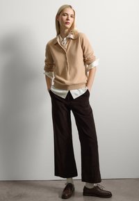 Beige gestrickte Strickjacke über einem weißen Hemd, kombiniert mit braunen weiten Hosen. Braune Schuhe vervollständigen das Outfit, mit sichtbaren weißen Socken.