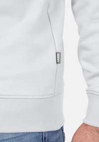 Sweatshirt gris clair en coton doux avec des poignets et un ourlet côtelés. Présente une petite étiquette logo grise sur le côté. Coutures détaillées visibles.