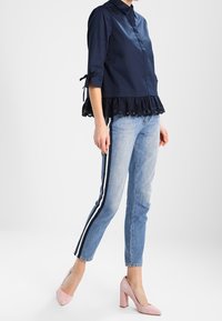 Marinblus med knäppning framtill och 3/4-ärmar, prydd med spetsdetaljer vid nederkanten. Buren tillsammans med ljusblå jeans och blekrosa klackar.