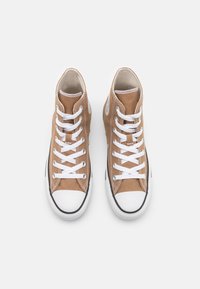 Para beżowych trampków typu high-top z płótna, z białymi gumowymi noskami, białymi sznurówkami i czarnymi wykończeniami, widziana z góry na białym tle.
