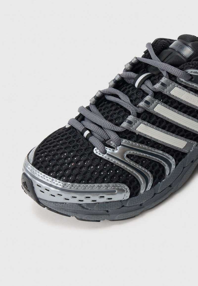 Scarpa da ginnastica nera con tomaia in mesh, dettagli metallici grigi e suola testurizzata per una migliore aderenza. I lacci sono grigi con una protezione sicura sulla punta.