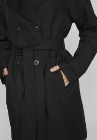 Juodas dvigubas paltas su diržu juosmenyje, turintis dvi priekinės kišenes ir apvalius juodus mygtukus. Lygus, minkštas audinys su pritaikytu siluetu.