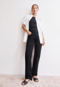 Jean noir évasé associé à un haut noir ajusté et une chemise blanche surdimensionnée, portée avec des sandales à talons noires avec des accents texturés.