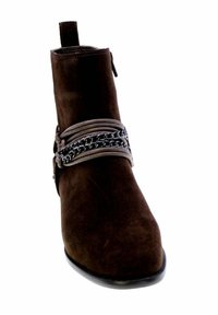 Stivaletto in suede marrone con punta affusolata, accentuato da cinghie sovrapposte e un dettaglio in catena argentata. Caratterizzato da un taglio per il rilascio e una cerniera laterale.