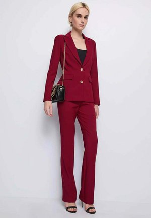 Donna in piedi contro il muro che indossa un tailleur pantalone color bordeaux su misura, top nero, sandali neri con tacco e borsa a tracolla nera con catenina dorata.