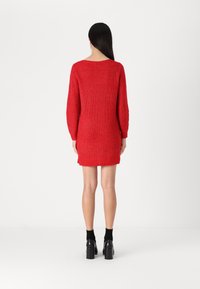 Una modella sta in piedi con la schiena rivolta verso la telecamera, indossando un aderente vestito a maglia rosso e stivali neri con tacco su uno sfondo bianco.