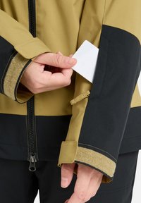 Gelb und schwarze Jacke mit strukturiertem Bündchen und Reißverschluss. Eine Hand steckt eine weiße Karte in eine Seitentasche. Verstellbares Bündchendetail sichtbar.