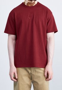 Maglietta in cotone color bordeaux con collo a giro e maniche corte. Presenta un logo Nike ricamato sul petto. Tessuto strutturato. Design semplice.