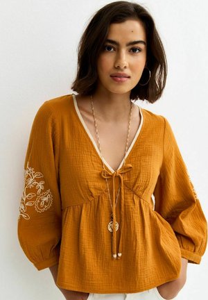Młoda kobieta o ciemnych włosach do ramion, ubrana w musztardowożółtą bluzkę z haftowanymi rękawami i długi złoty naszyjnik.