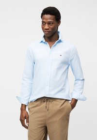 Άνδρας που φοράει ανοιχτό μπλε πουκάμισο με κουμπιά και μπεζ παντελόνι με κορδόνι στη μέση, στέκεται με το ένα χέρι στην τσέπη, κοιτώντας πλάγια με ελαφρύ χαμόγελο.