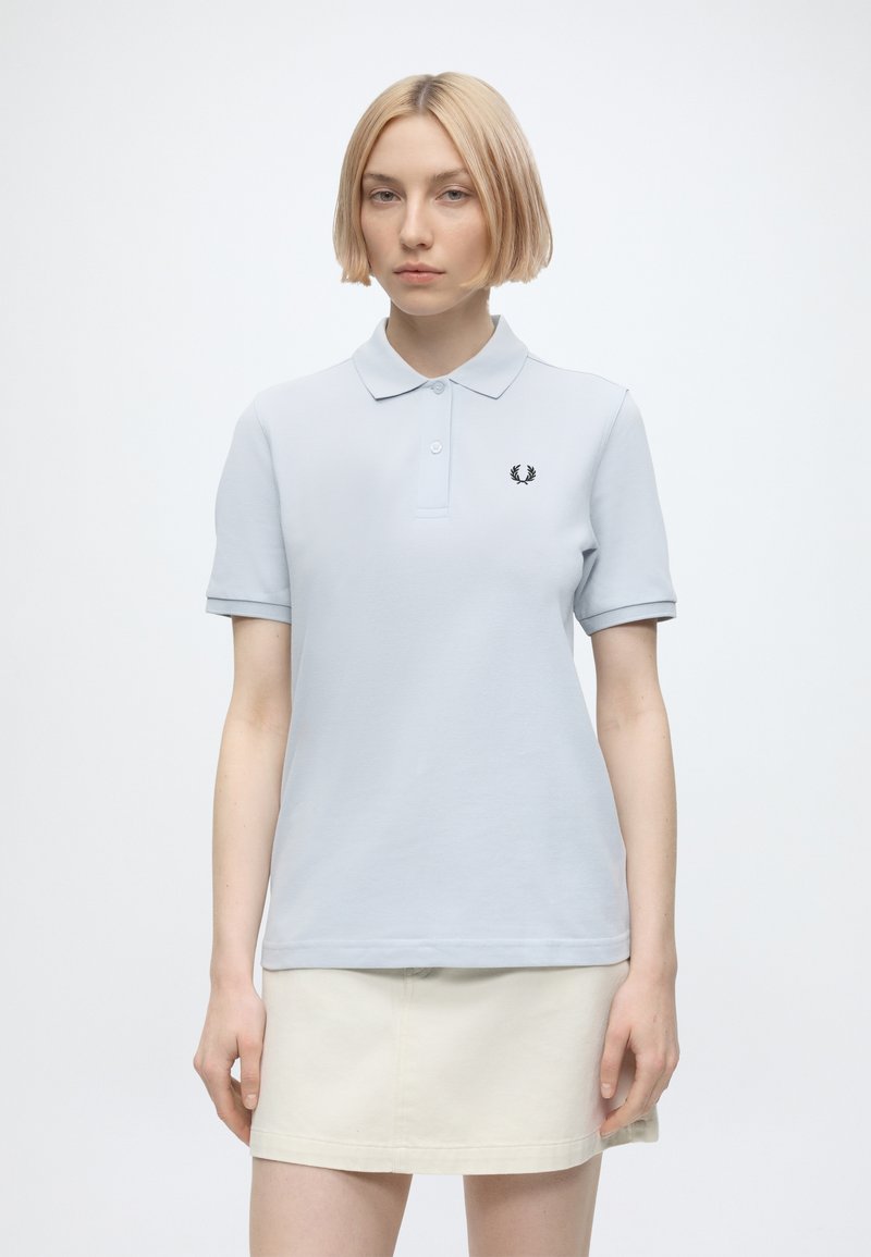 Jeune femme aux cheveux blonds courts portant un polo bleu clair à manches courtes et une jupe crème, debout devant un fond blanc uni.