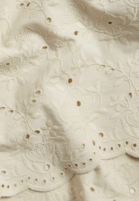 Broderad tyg i krämfärg med blommönster och utskurna detaljer. Texturen har upphöjd sömnad och scallopade kanter.